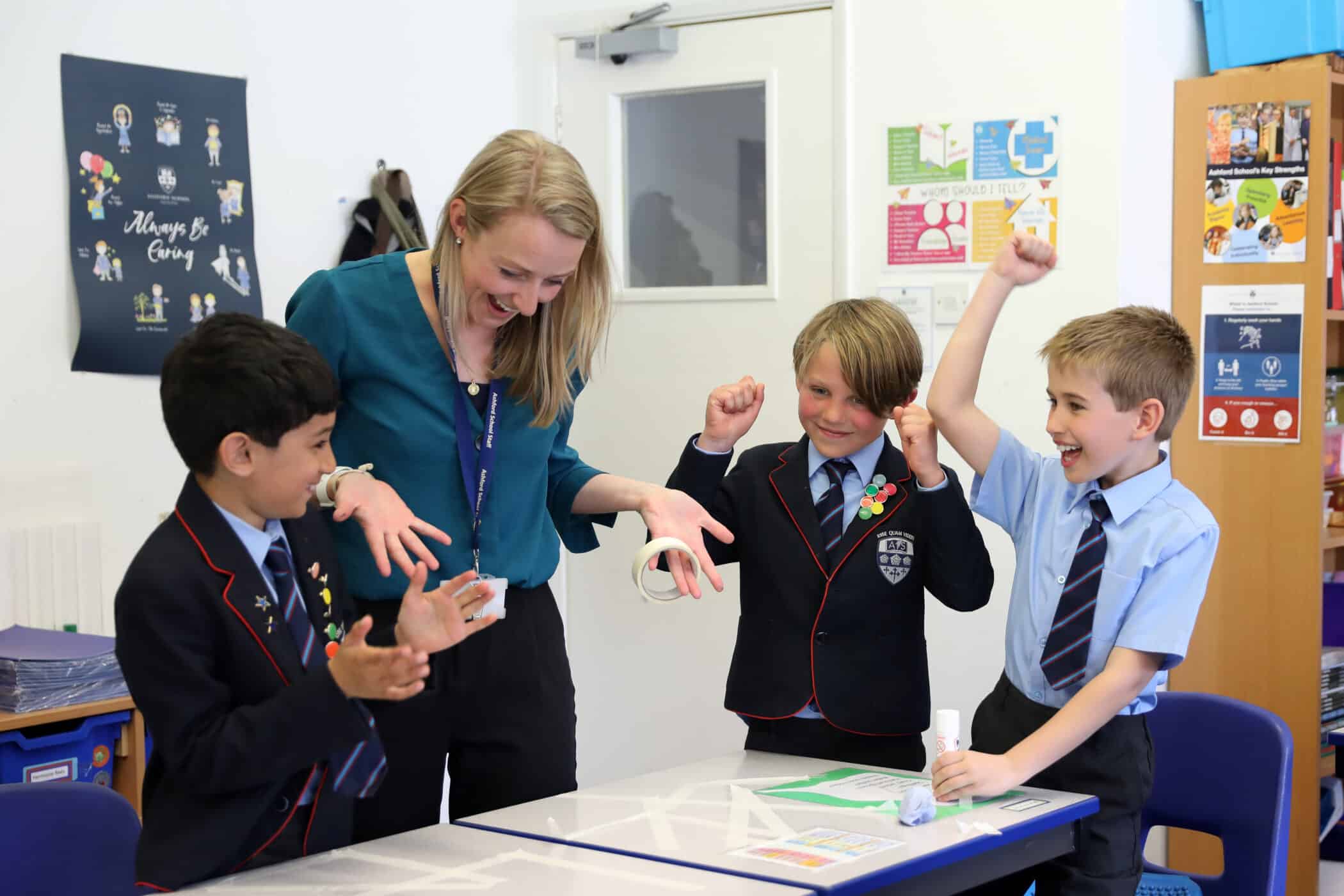 A teacher leans over a classroom table with three young boys in school uniform; the boys smile and raise their fists in celebration as they look at a project or work on the table.