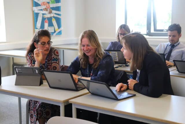 A group of teachers sit at desks in a bright classroom, working together on laptops and tablets; three women in the foreground discuss something on their screens while other colleagues work in the background.