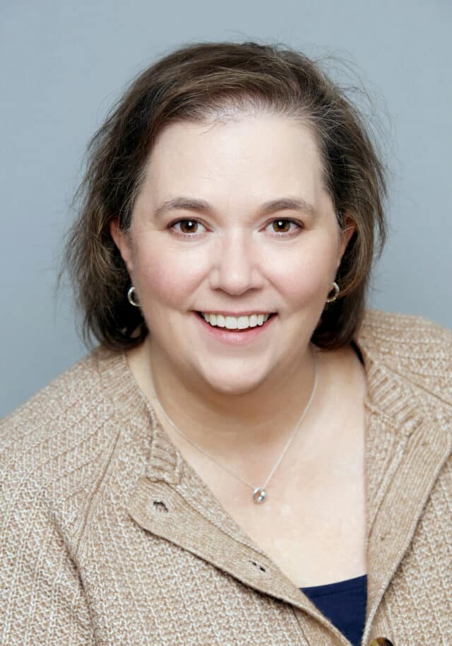 Woman smiling in a studio portrait, wearing a beige knit sweater and silver necklace, with shoulder-length brown hair and a neutral grey background.