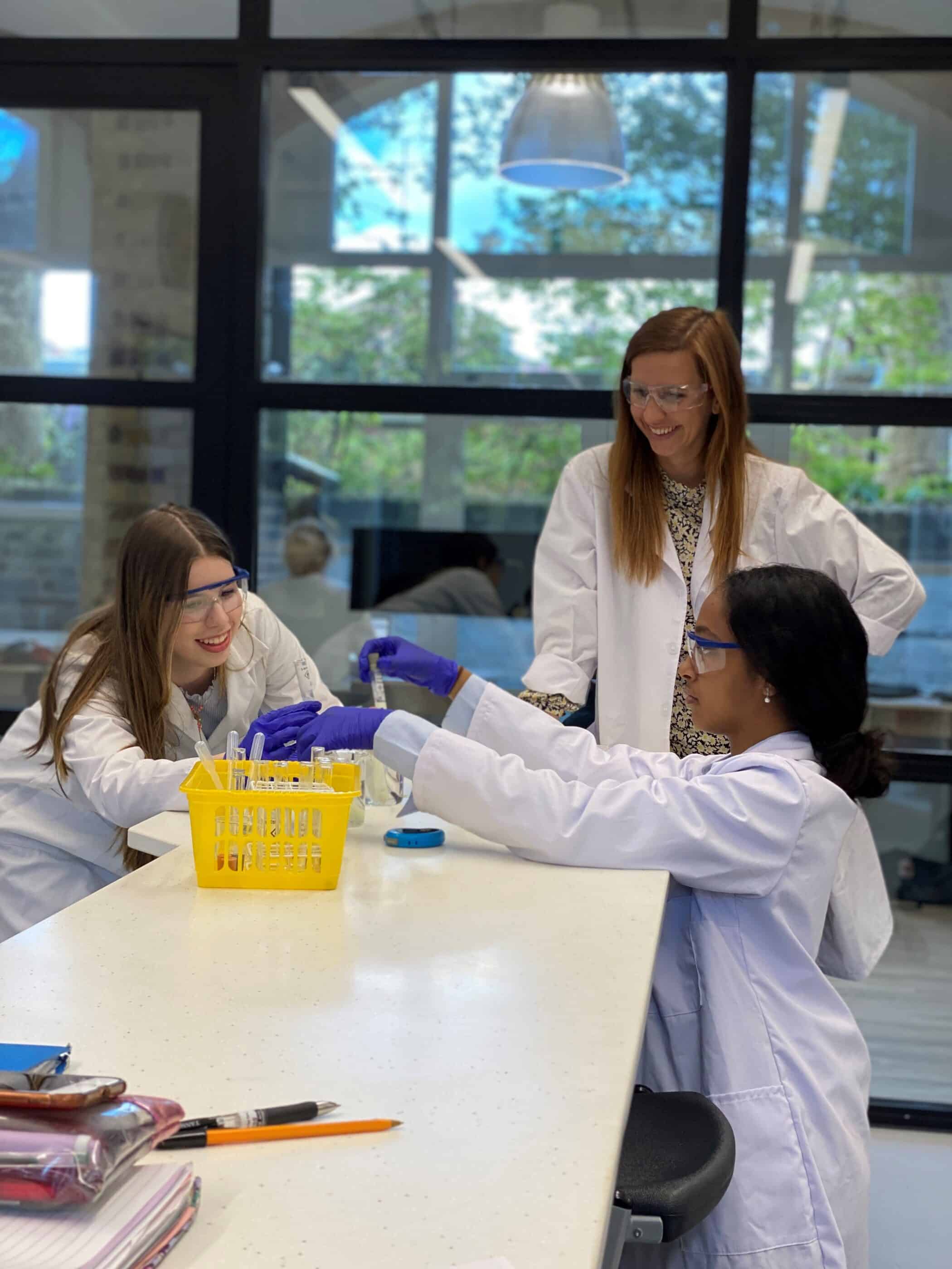 Three students wearing white lab coats sit around a table conducting a hands-on science activity with pipettes and containers, while an adult supervisor in a lab coat stands behind them, smiling and observing in a bright classroom or laboratory setting.
