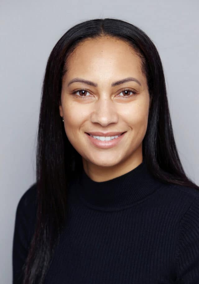 Woman with long dark hair smiling, dressed in a black ribbed top, photographed against a light grey background.