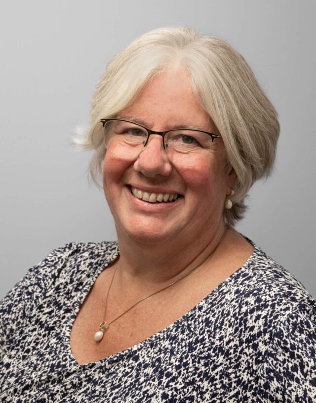 A smiling woman with short, silver-grey hair and glasses, wearing a navy and white patterned top, pearl earrings and a pendant necklace. She faces the camera against a plain grey background.