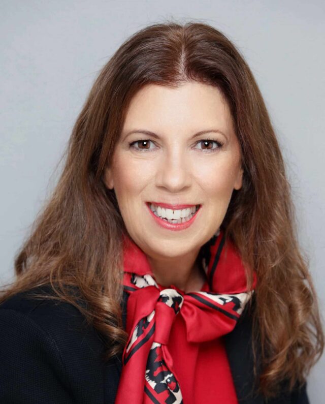 Woman smiling, wearing a red scarf with a patterned border over a black blazer, posed in front of a grey background.