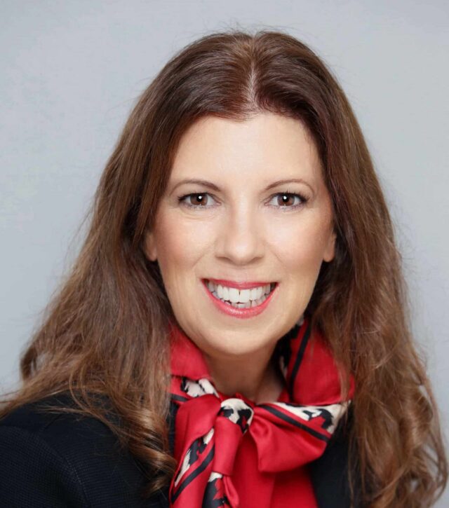 Woman smiling, wearing a red scarf with a patterned border over a black blazer, posed in front of a grey background.