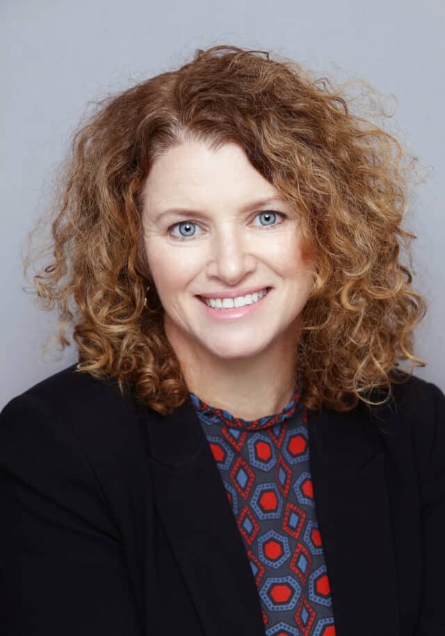 Woman smiling at the camera, with curly auburn hair, wearing a patterned blue and red top under a black blazer, posed against a plain light grey background.