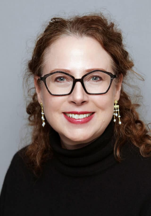 Woman with glasses and earrings smiling, wearing a black top and red lipstick, set against a plain light grey backdrop.
