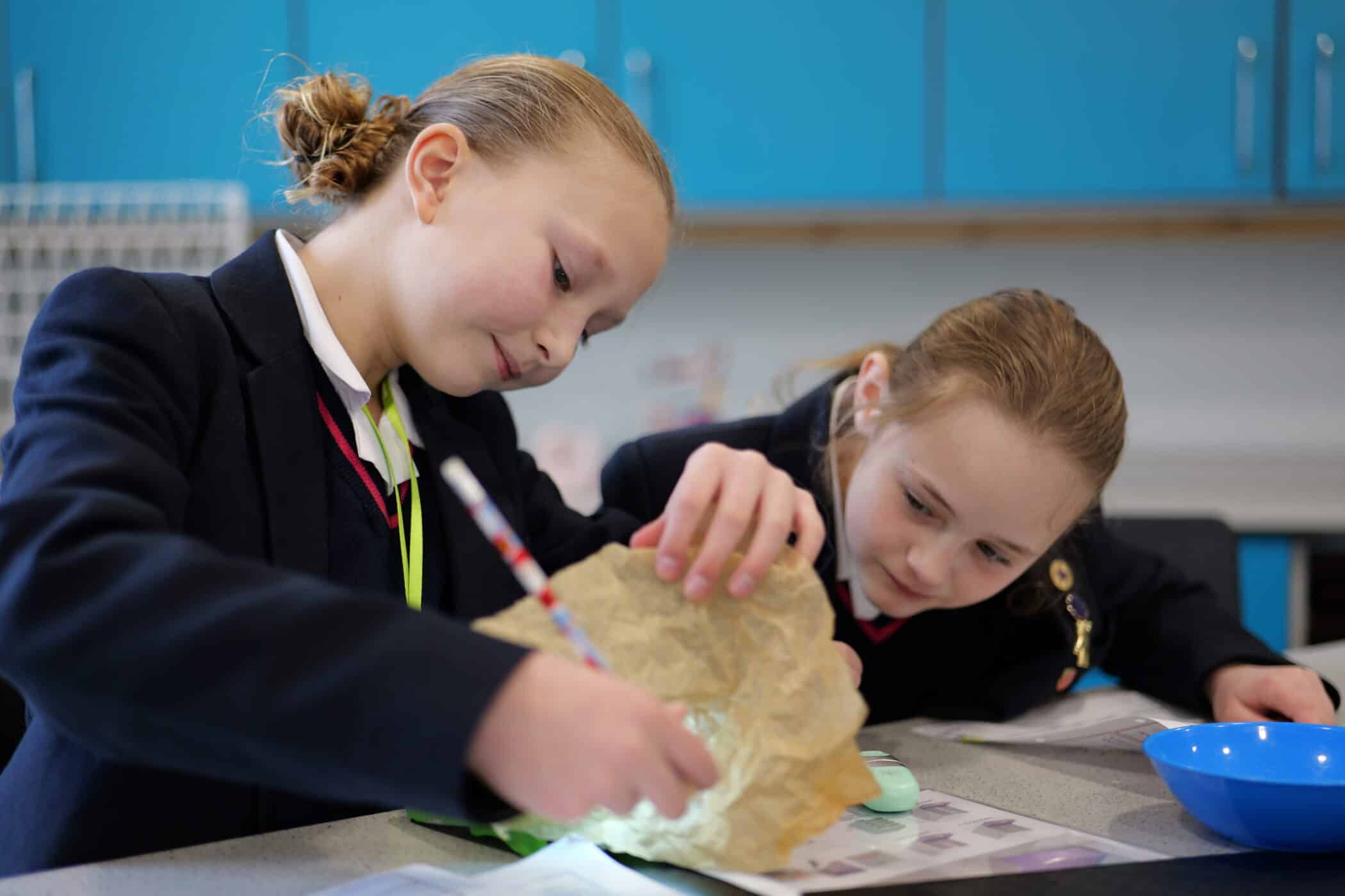Two pupils in school uniform lean over a table, closely examining an object inside a paper covering. One uses a pencil while both focus intently on the task.