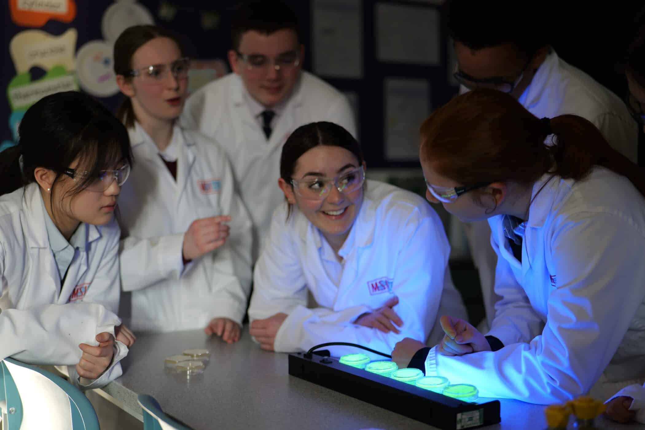 A group of students in lab coats and safety glasses gather around a bench in a darkened science room. They watch glowing samples under a blue light, smiling and discussing the experiment.