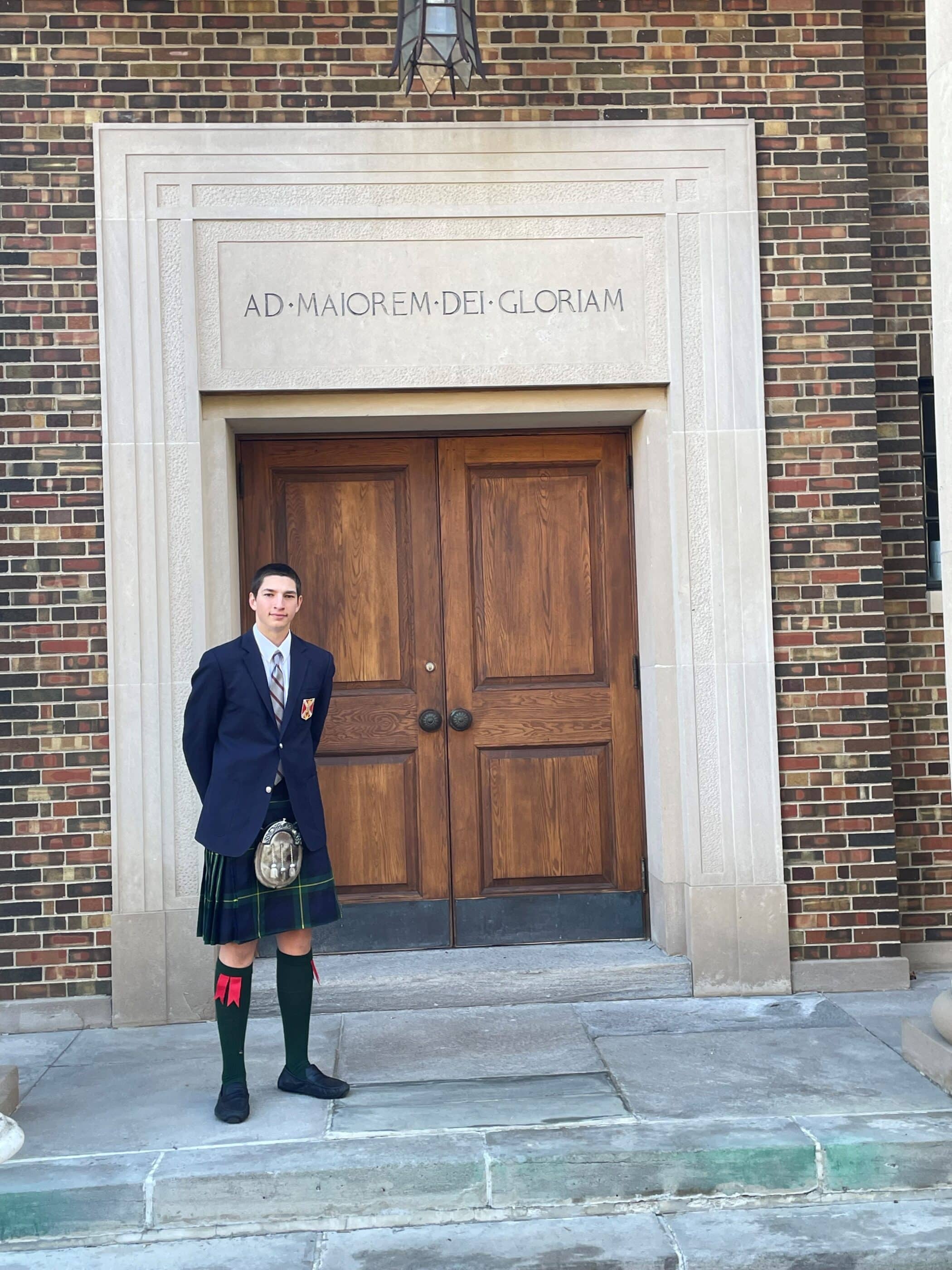 Para12Biff.jpg Enter value here Enter value here Young man in a school blazer and tartan kilt stands in front of a large wooden doorway framed by stone, with the Latin inscription “Ad Maiorem Dei Gloriam” engraved above.