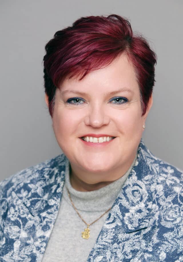 Woman smiling, with short dark red hair, wearing a light blue patterned blazer and a gold necklace, on a neutral background.