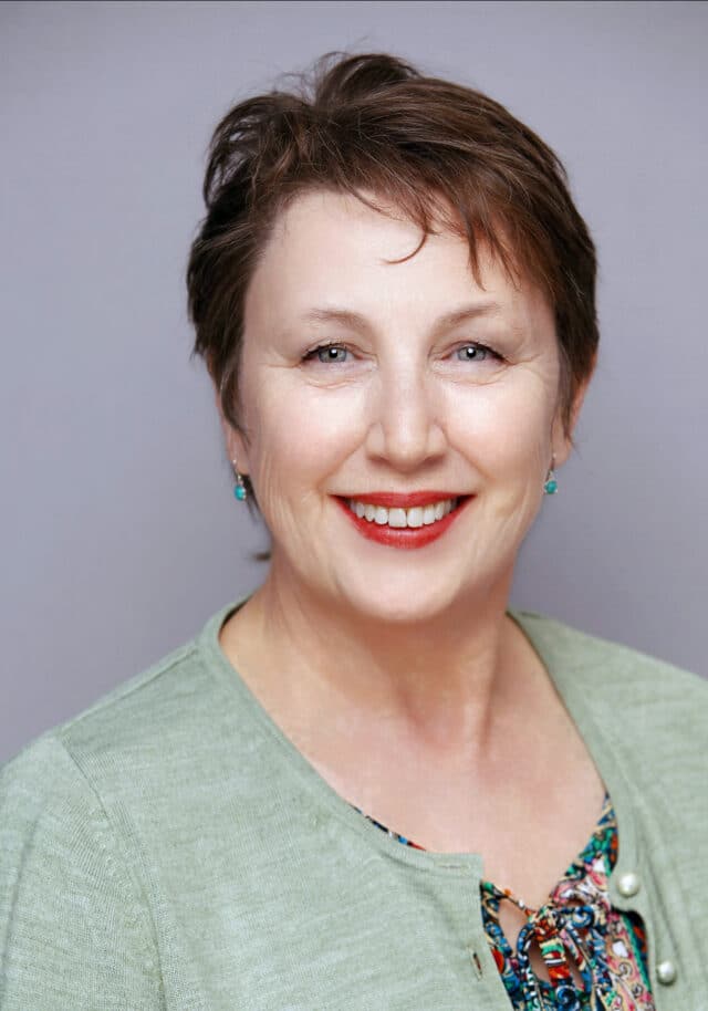 Woman with short hair smiling, wearing a light green cardigan over a patterned top, posed in front of a grey background.