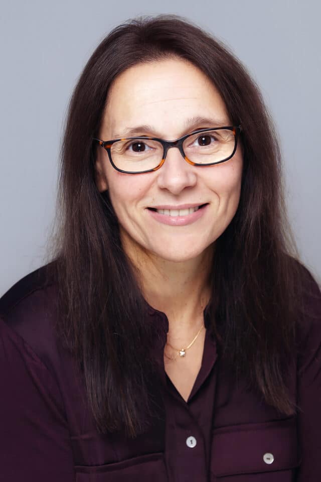 Woman with dark brown hair smiling, wearing tortoiseshell glasses and a deep purple blouse, posed against a neutral grey background.