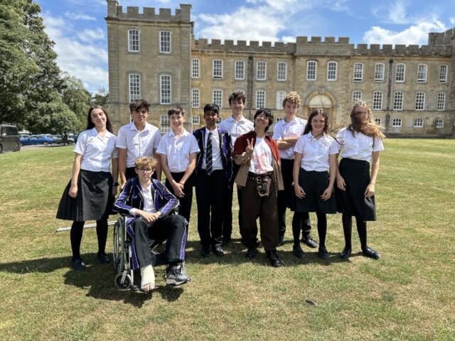 A group of nine students in school uniforms and one casually dressed adult posing together on a lawn in front of a large historic stone building. One student is seated in a wheelchair at the front, while the others stand behind, smiling in bright sunny weather.