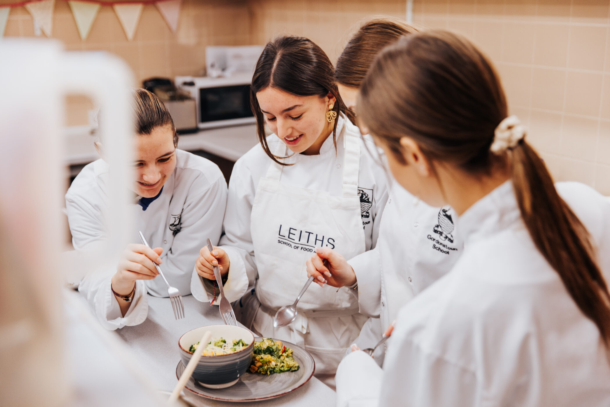 Four students wearing white chef uniforms and aprons stand around a kitchen counter, tasting and evaluating a plated dish together. One apron reads “Leiths School of Food and Wine,” and they hold forks and spoons as they smile and focus on the food in a professional training kitchen.
