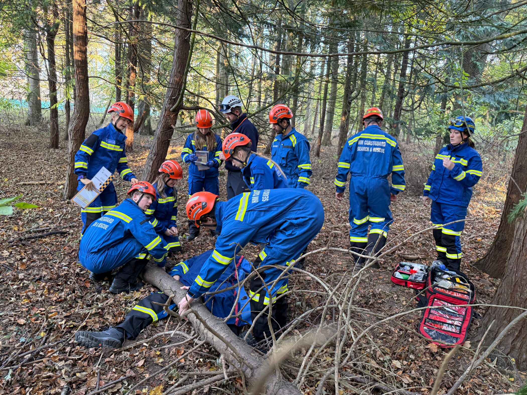 A team of search and rescue trainees in blue uniforms and orange helmets kneel in a wooded area, practising a first aid scenario on a person lying on the ground. A fallen tree branch and medical bags are visible among the leaves.