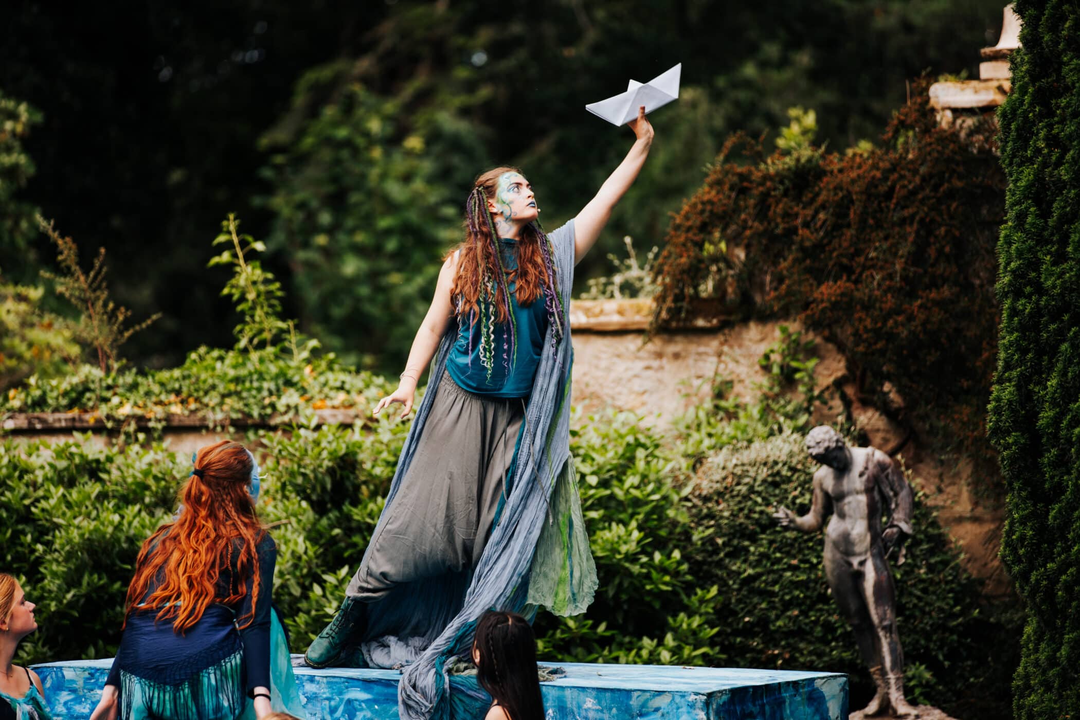 An outdoor theatre performance of The Tempest, with a young actor standing on a platform holding a paper boat aloft. She wears flowing, layered costumes in blue and green tones, while other performers sit nearby in a garden setting with statues and greenery in the background.