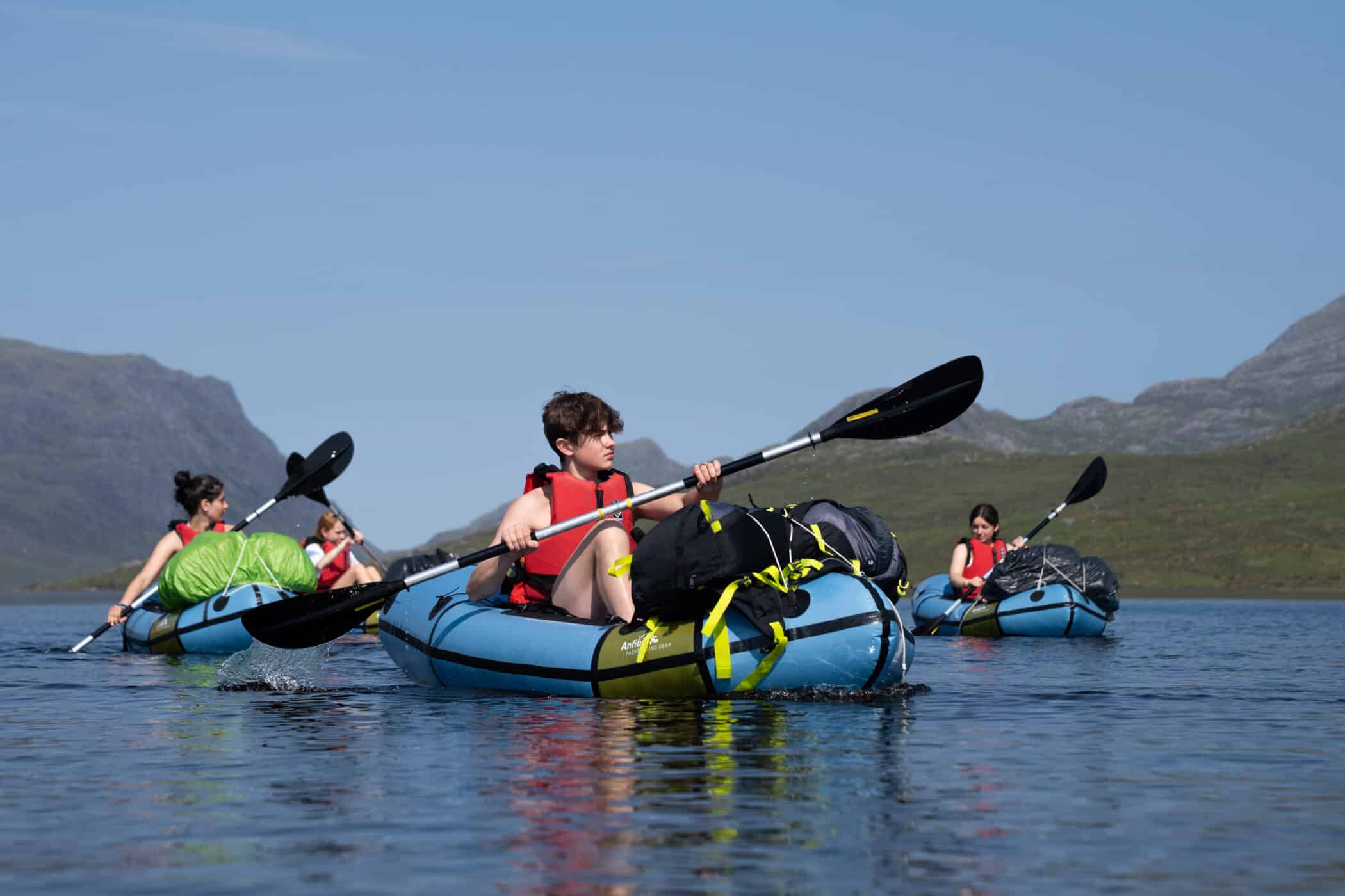 Four teenagers wearing life jackets paddle inflatable kayaks across a calm lake, surrounded by open water and rolling hills under a clear sky.