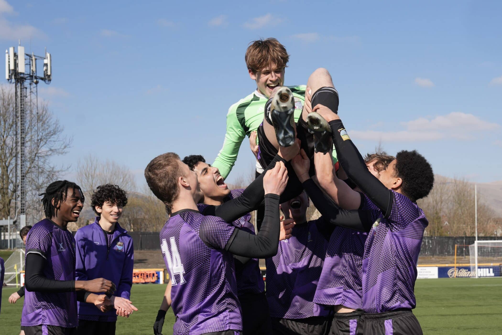 A football team in purple kits celebrate on the pitch by lifting a teammate into the air, smiling and cheering after their victory, with a goal and hills visible in the background.