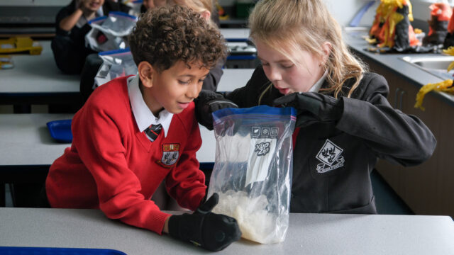 Two primary-aged pupils in school uniform and protective gloves examine a clear zip bag filled with ice during a classroom science activity.