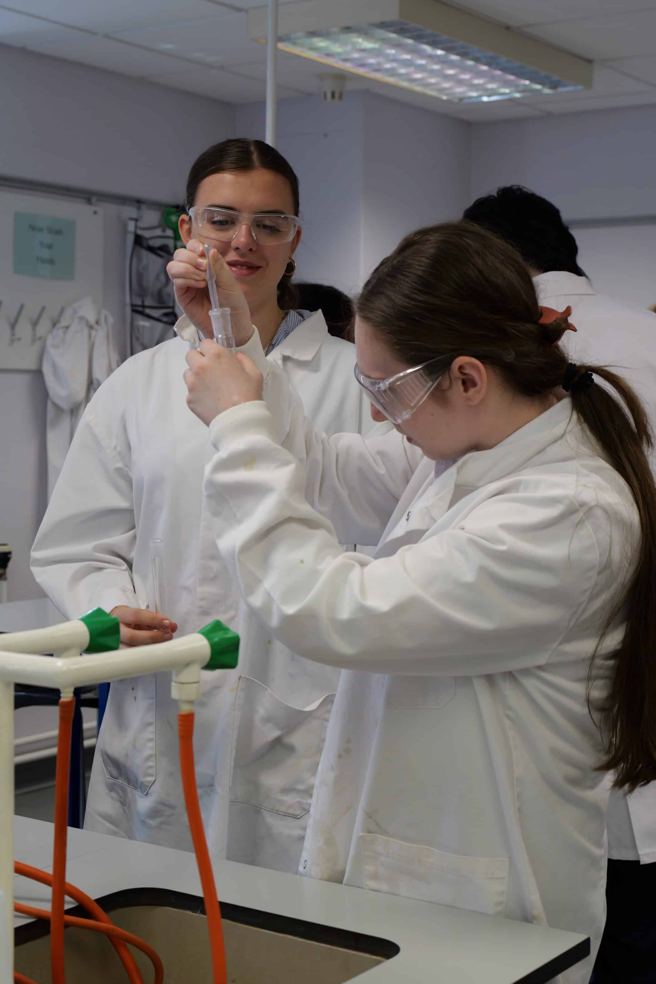 Two students wearing safety goggles and white lab coats conduct an experiment in a science laboratory. One carefully uses a pipette to transfer liquid into a test tube while the other observes closely beside a lab bench with taps and equipment.