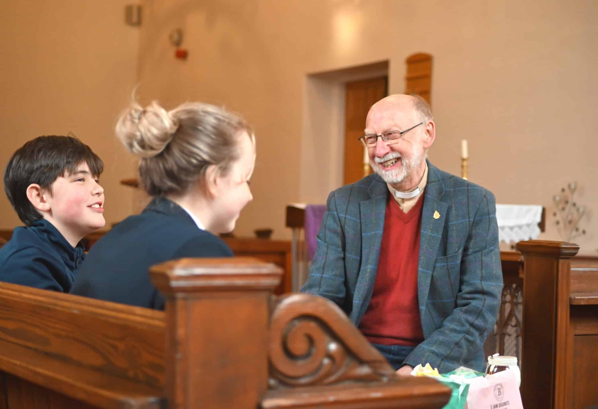 An older man wearing glasses, a tweed jacket, and a clerical collar sits on a wooden church pew, smiling and laughing as he talks with two children seated beside him. The children, both in dark school uniforms, face him and smile. The setting appears to be a church interior, with wooden pews, candles, and an altar visible in the background.