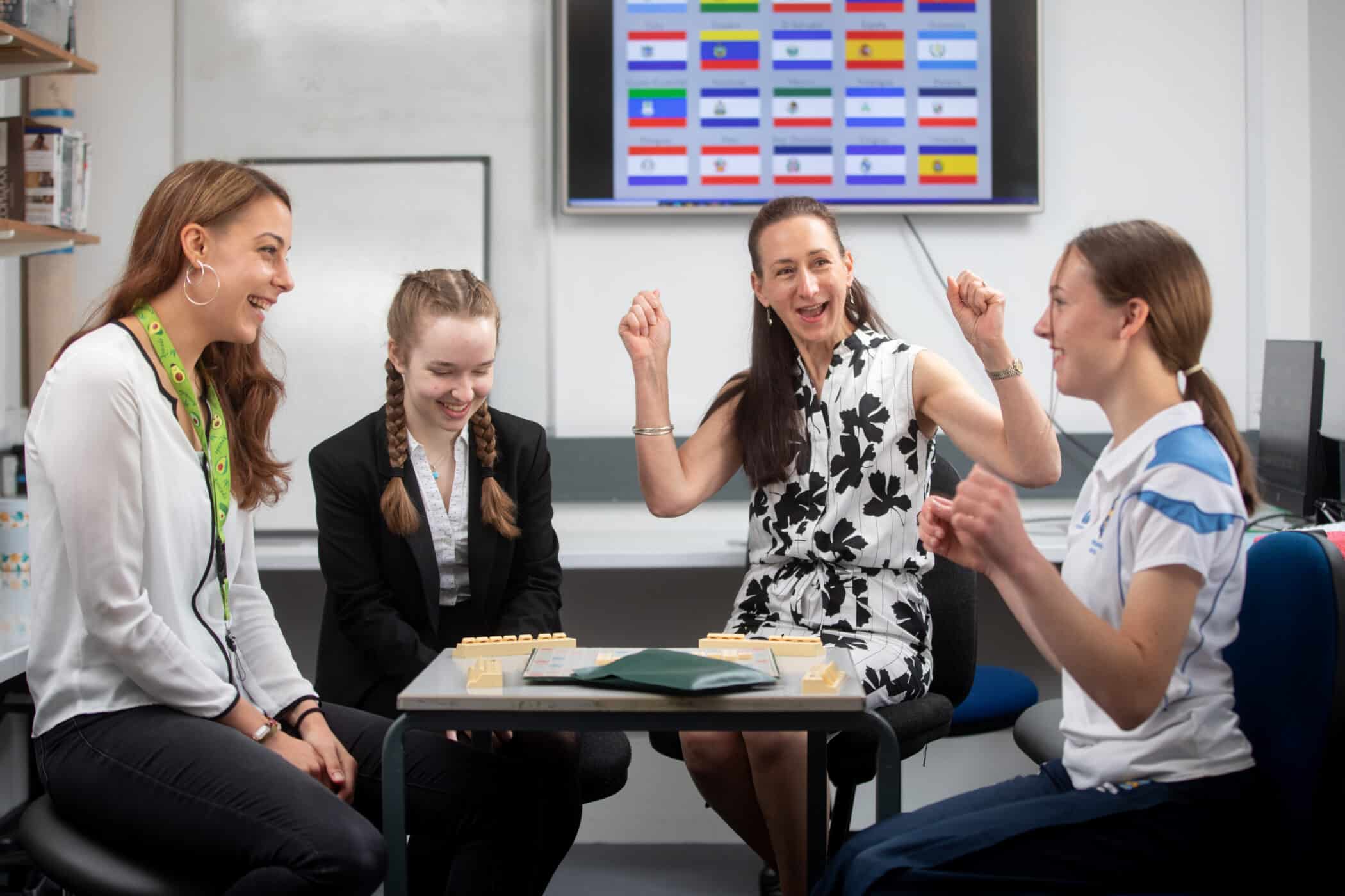 Four people sit around a small table in a classroom or office setting, smiling and laughing during a tabletop game. A woman in a sleeveless black-and-white dress raises her arms in celebration, while the others—two young women and one adult woman—look on and smile. Game pieces are arranged on the table, and a screen displaying rows of national flags is visible on the wall behind them.