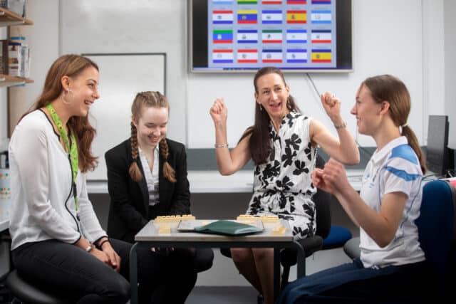 Four people sit around a small table in a classroom or office setting, smiling and laughing during a tabletop game. A woman in a sleeveless black-and-white dress raises her arms in celebration, while the others—two young women and one adult woman—look on and smile. Game pieces are arranged on the table, and a screen displaying rows of national flags is visible on the wall behind them.