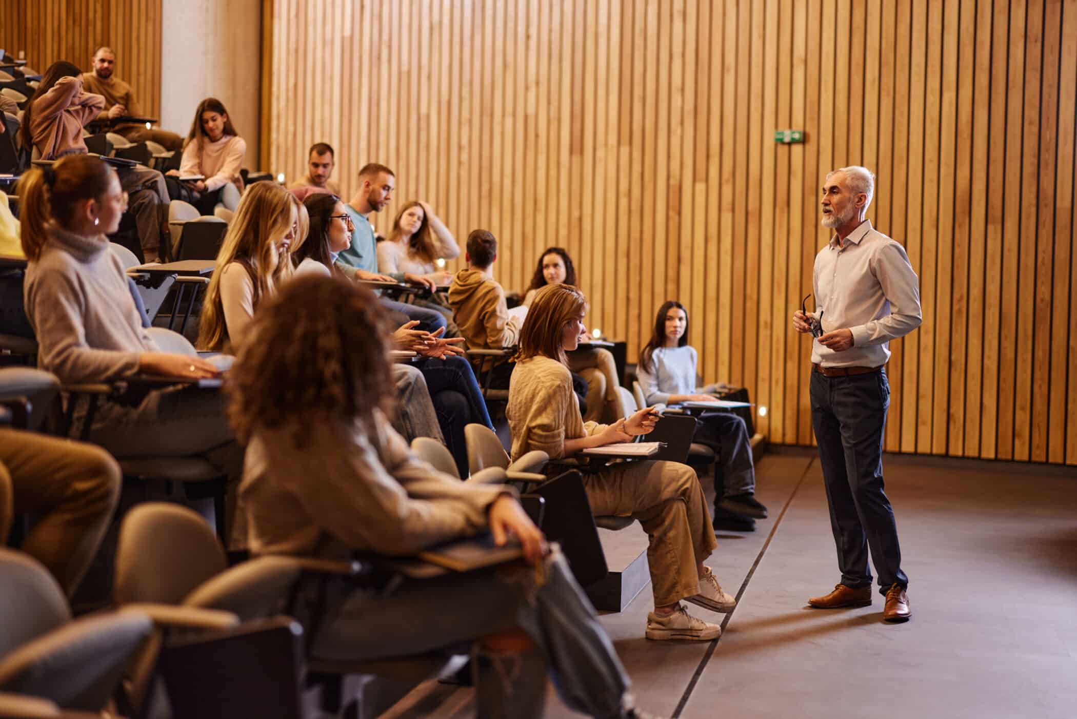 A university lecturer stands at the front of a tiered lecture hall, speaking to a group of students seated in rows. The lecturer, an older man with gray hair and a beard, holds a pair of glasses while addressing the class. Students of various genders sit with notebooks and laptops, listening and taking notes. The room features wooden panel walls and auditorium-style seating.