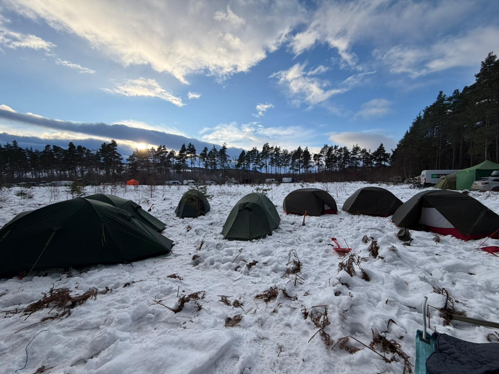 Several small camping tents arranged across a snowy clearing surrounded by trees, with the sun low on the horizon and vehicles and larger tents visible in the distance.