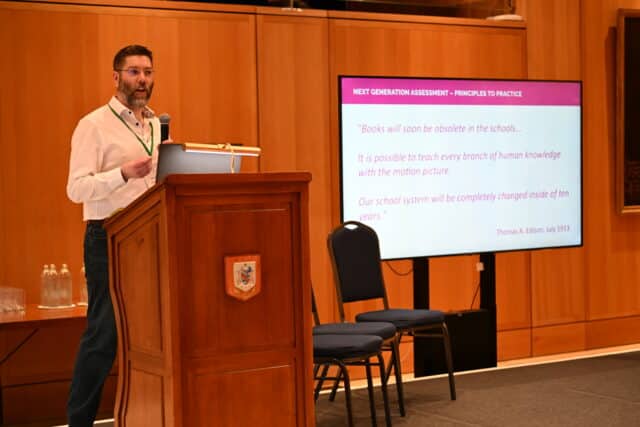 Man speaking at a wooden podium in a conference room, holding a microphone and presenting a slide titled “Next Generation Assessment – Principles to Practice,” displaying a 1913 quote by Thomas Edison about books becoming obsolete in schools; chairs and a large screen behind him.