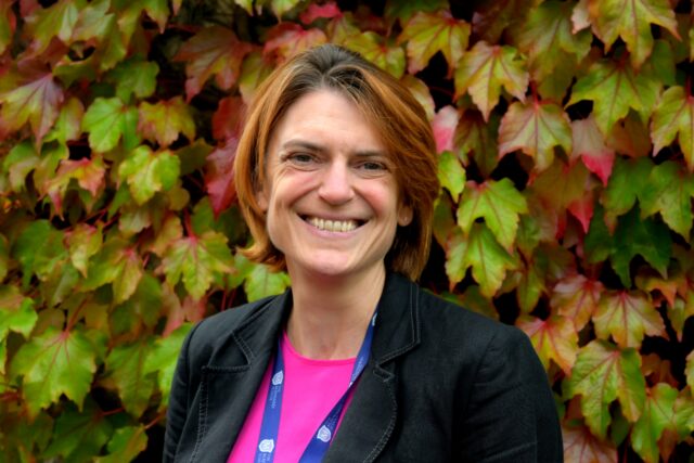 A smiling woman with short, light brown hair stands outdoors in front of a wall covered in colorful autumn leaves. She is wearing a black blazer over a bright pink top and a lanyard around her neck.