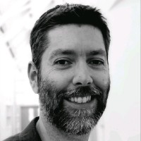 Black and white headshot of a smiling man with short dark hair and a full beard, facing the camera against a softly blurred indoor background.