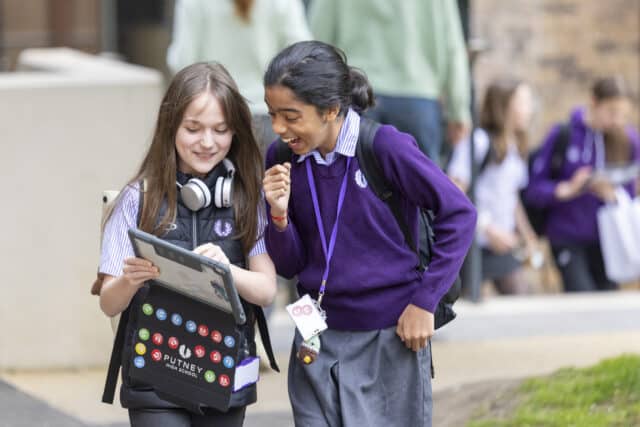 Two students in purple school uniform stand outdoors, looking at a tablet and smiling, with other pupils blurred in the background.