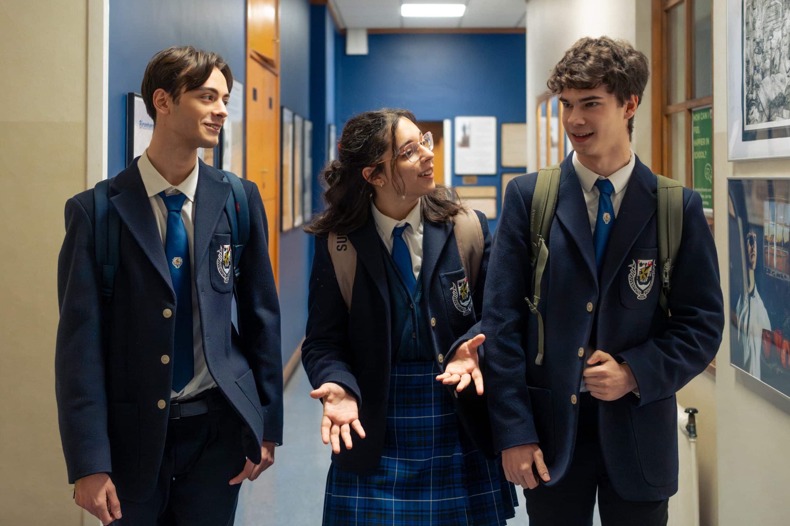 Three students in school uniforms walk down a brightly lit school hallway while talking and smiling. The student in the middle gestures with her hands as she speaks, while the two students on either side listen and look at her. All three wear navy blazers, blue ties, and backpacks, and framed photos and notices line the corridor walls.
