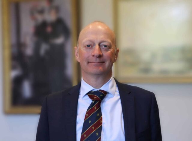 A middle-aged man with a bald head and light skin stands facing the camera, wearing a dark suit jacket, light blue dress shirt, and a striped tie in red, green, and gold. He has a slight smile and a calm expression. The background is softly blurred, showing framed artwork on a wall.