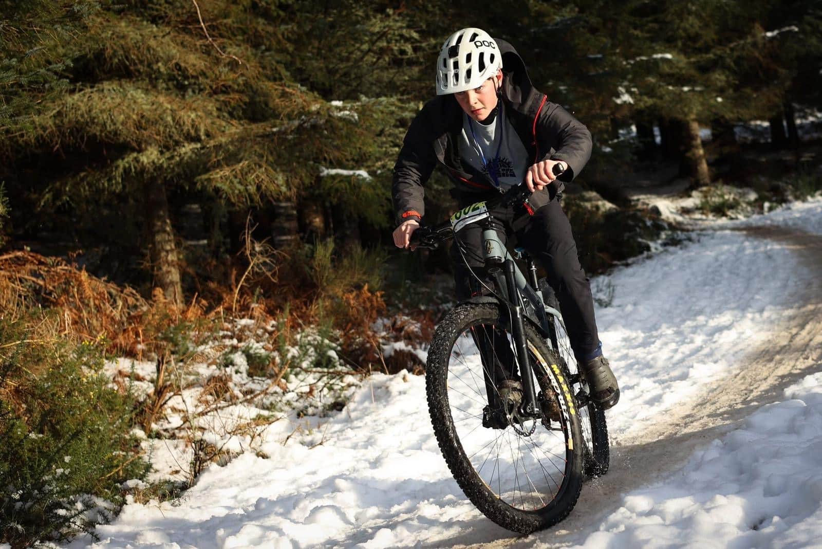 A mountain biker wearing a helmet rides along a snowy forest trail, leaning into a turn while kicking up snow.