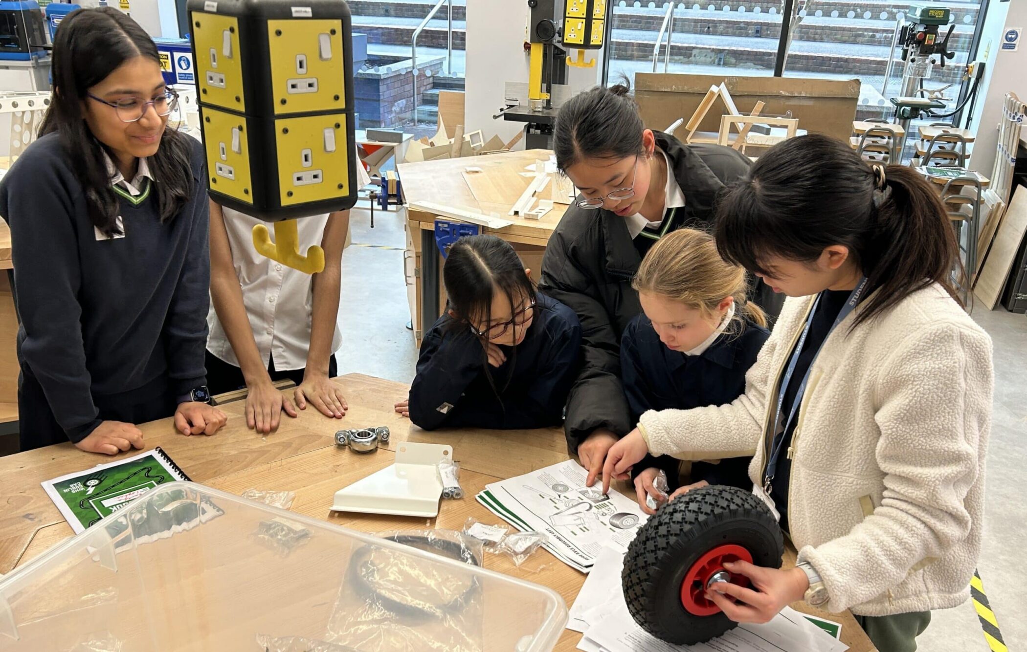 A group of students in a school workshop gather around a table assembling a large wheel using tools and instruction sheets. One student holds the wheel while others examine the instructions, with mechanical parts and wiring kits spread across the table.