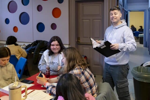 A group of young people sit around a table in a classroom or workshop space while an instructor stands nearby speaking and holding a clipboard. Papers, phones, and drinks are spread across the table, and colourful circular wall panels decorate the room.
