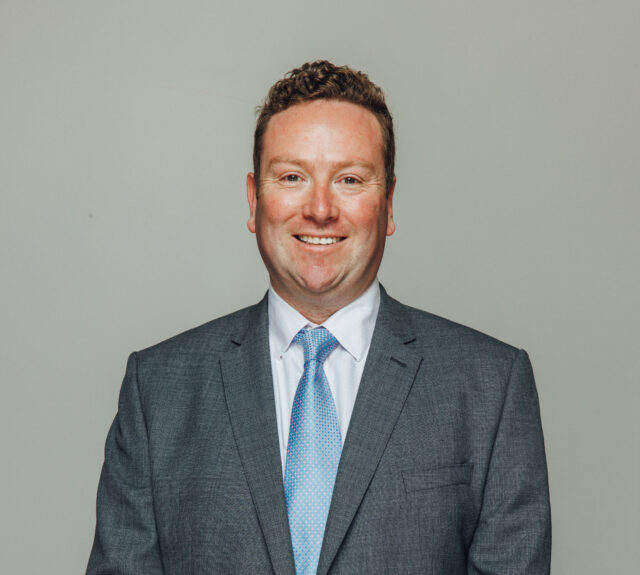 A man stands facing the camera against a plain light-colored background, smiling warmly. He is wearing a grey suit jacket with a white dress shirt and a light blue patterned tie. His hands are clasped in front of him, and he appears neatly groomed with short, curly hair.