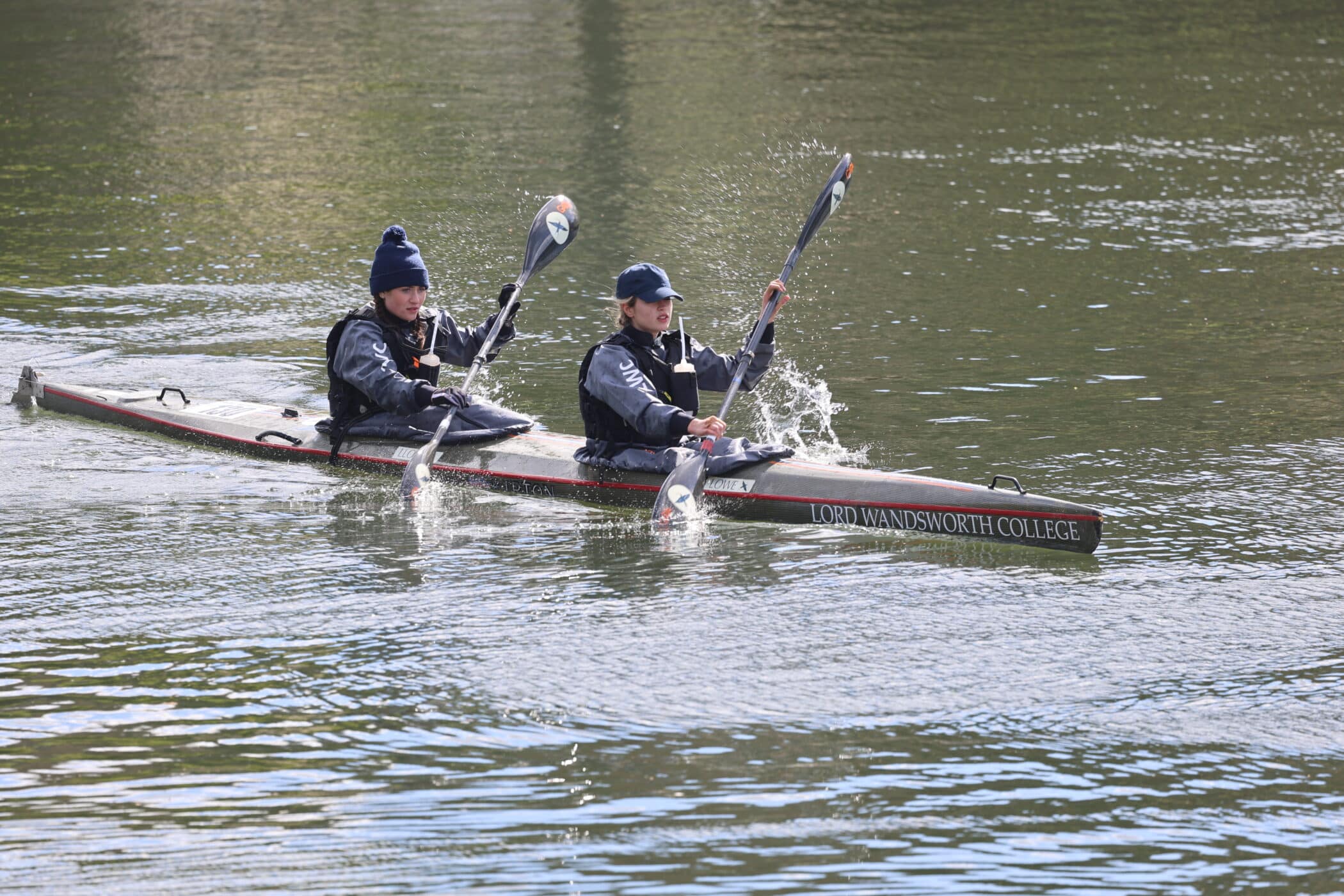 Two paddlers in a canoe stroke in sync on a river, splashing water as they paddle, both wearing dark clothing and hats.