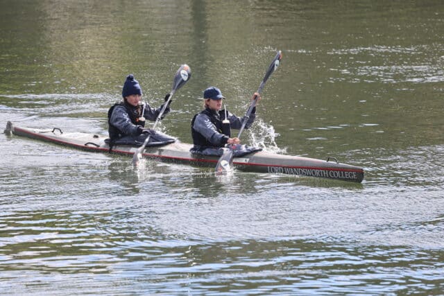 Two paddlers in a canoe stroke in sync on a river, splashing water as they paddle, both wearing dark clothing and hats.