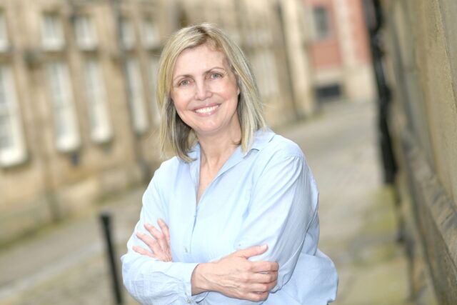 Smiling woman with blonde hair wearing a light blue shirt, standing outdoors with arms crossed in a blurred urban street setting.