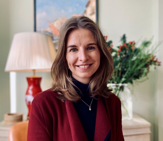 Smiling woman with shoulder-length brown hair wearing a burgundy jacket over a black top, seated indoors. Behind her are a table lamp, framed artwork and a vase of greenery, creating a warm, homely setting.