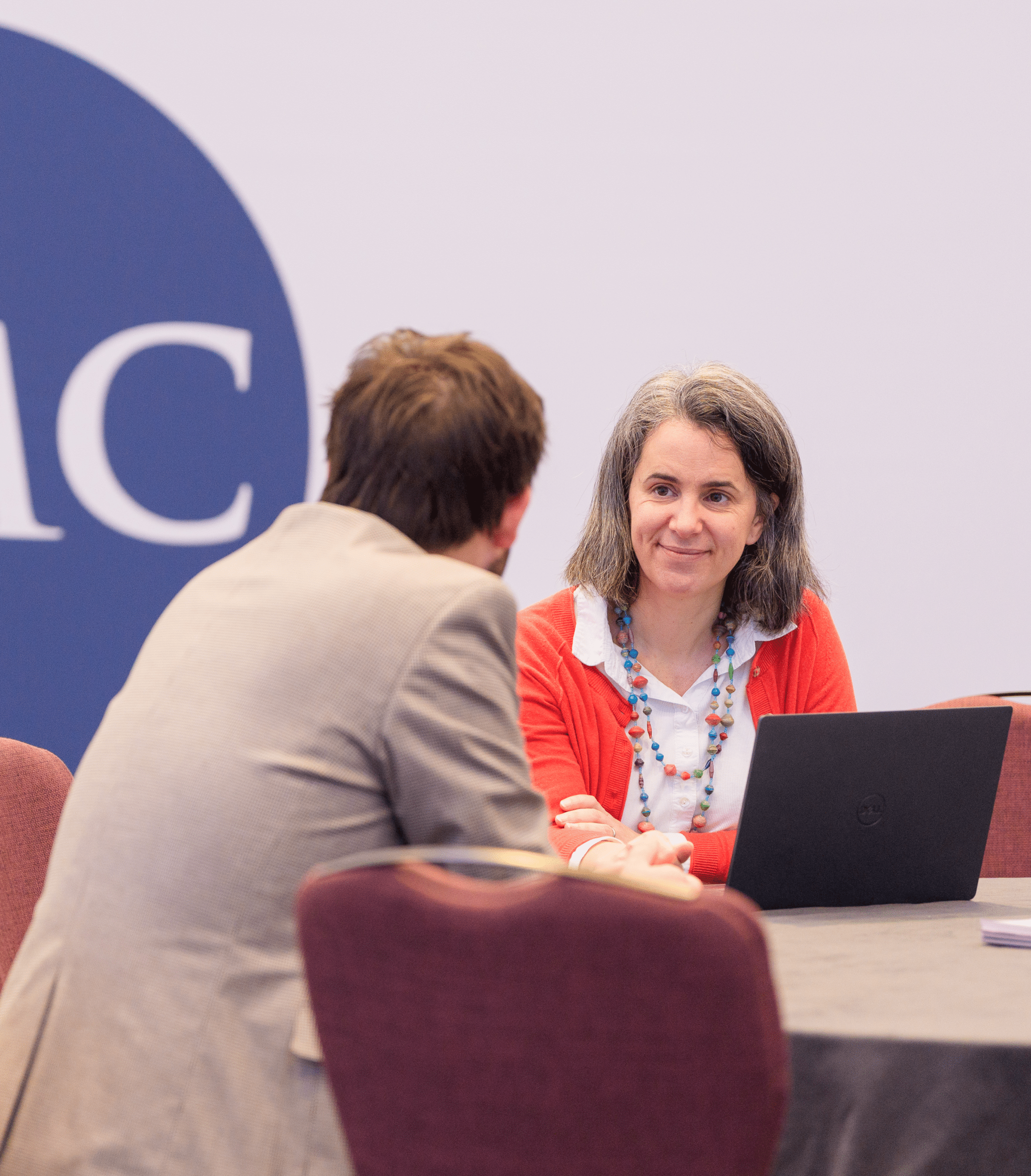 Two people seated at a table in a professional setting, engaged in conversation; a woman with a laptop smiles while facing a man, with a large “HMC” logo on the wall behind them.