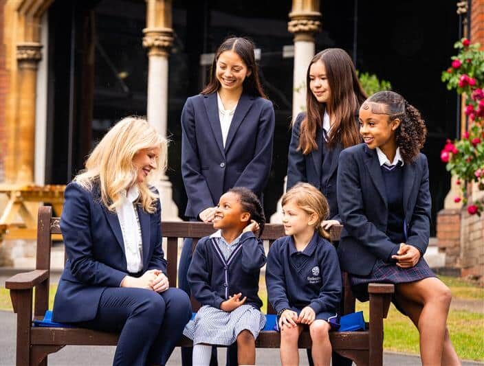School bench scene Woman in a suit sitting on a wooden bench outdoors, talking with a group of primary school children in school uniforms in a courtyard.