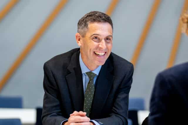 A smiling man in a suit and tie leans forward with his hands clasped, appearing engaged in conversation in a bright indoor setting.