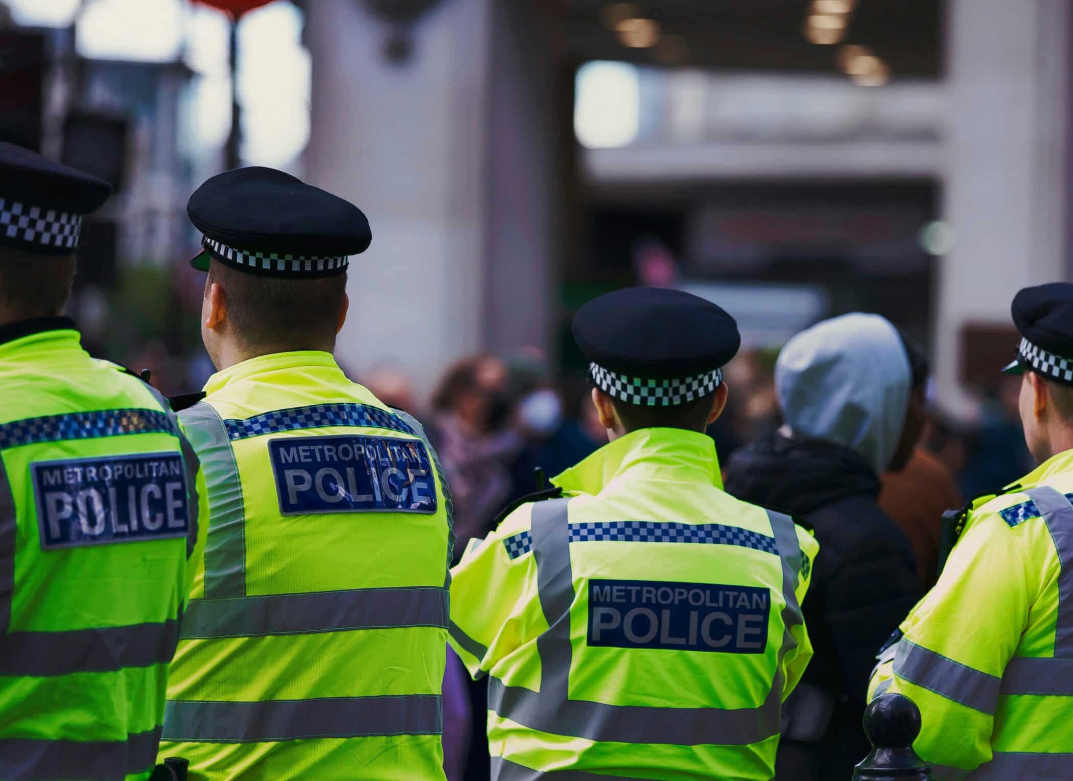 Four Metropolitan Police officers in high-visibility jackets standing with their backs to the camera, facing a blurred crowd in a city setting.