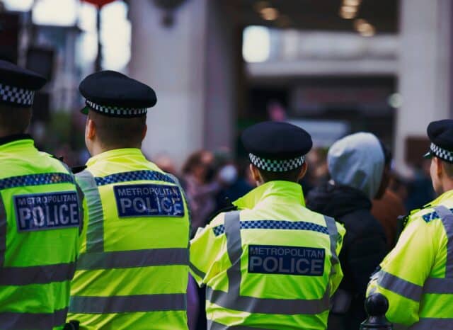 Four Metropolitan Police officers in high-visibility jackets standing with their backs to the camera, facing a blurred crowd in a city setting.