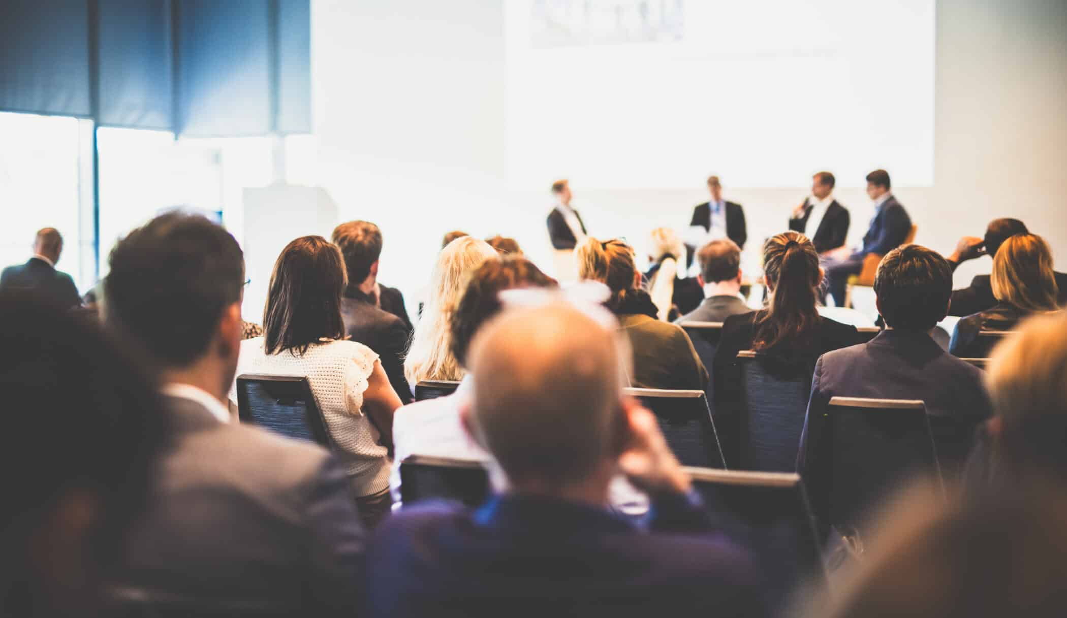 Audience seated in a conference room watching a panel discussion at the front, where several speakers are seated on stage. The image is softly focused, with attendees seen from behind and the panel slightly blurred in the distance.