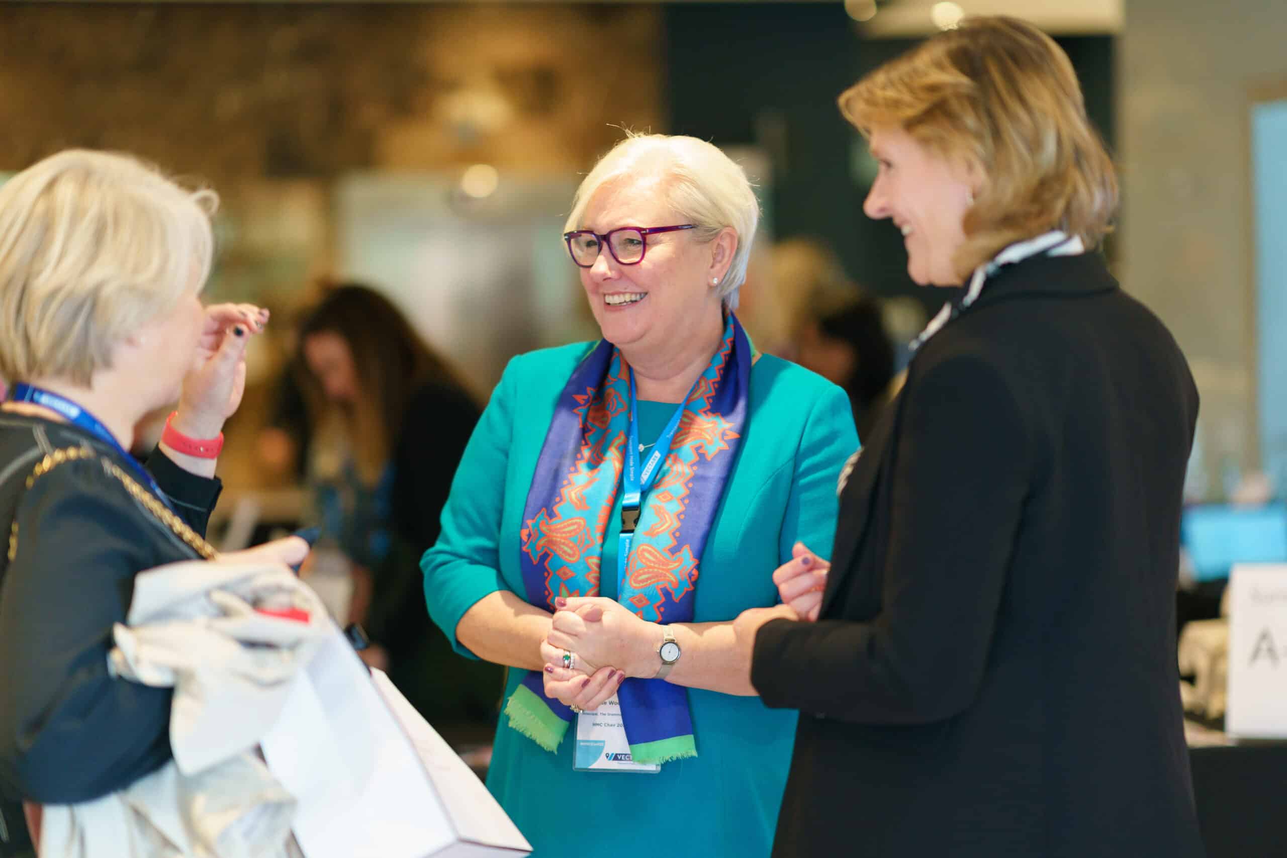 Three professionals chat and smile at a conference booth; central woman in teal dress and patterned scarf with glasses stands between two others, with blurred attendees and displays in the background.